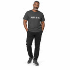 Full-body man wearing the asphalt JUST SI IT. t-shirt with white chest lettering and grey trainers, hands in trouser pockets on a white background.