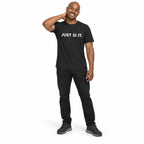 Full-body man wearing the dark grey JUST SI IT. t-shirt with white chest lettering and grey trainers, hand behind his neck on a white background.