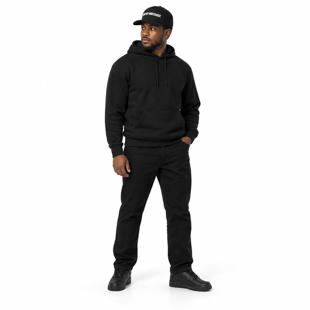 Full-body model wearing the black MAKE NO MISTAKES. embroidered cap with matching black hoodie and shoes, one hand in his pocket on a white background.