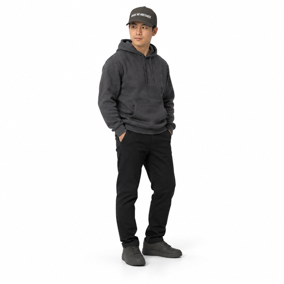 Full-body model wearing the dark grey MAKE NO MISTAKES. embroidered cap with a matching charcoal hoodie and shoes, hands in pockets on a white background.