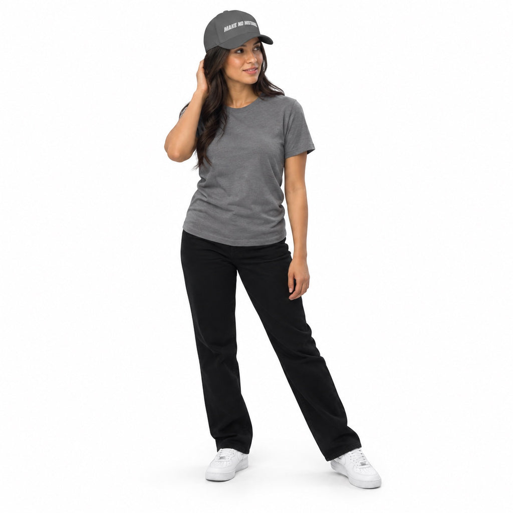 Full-body model wearing the grey MAKE NO MISTAKES. embroidered cap with a matching grey T-shirt and white shoes, touching her hair on a white background.