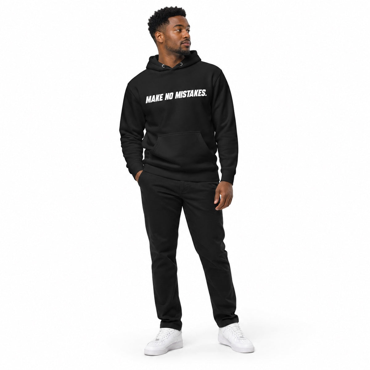 Full-body model wearing the black MAKE NO MISTAKES. hoodie with white chest print, one hand in his pocket and looking aside on a plain white background.