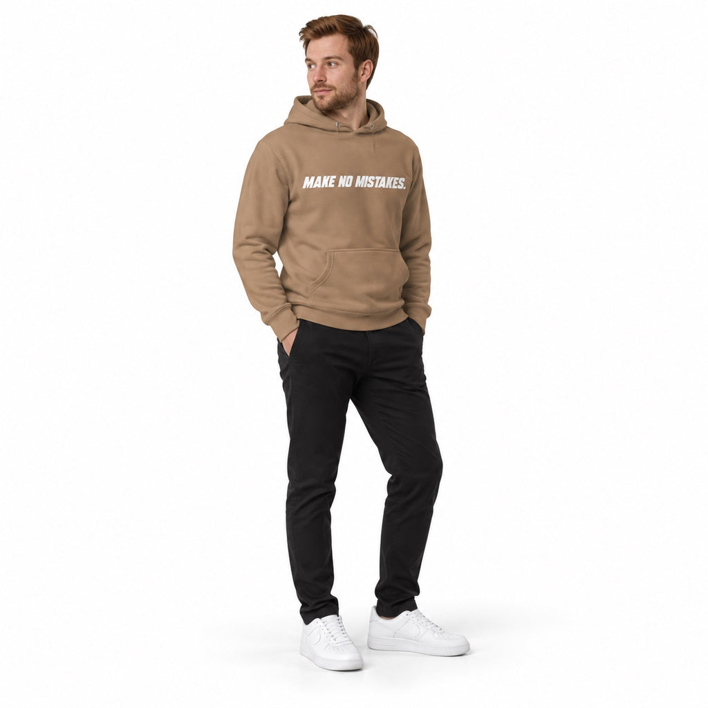 Full-body model wearing the latte brown MAKE NO MISTAKES. hoodie with white chest print, torso slightly turned and hands in his pockets on a plain white background.