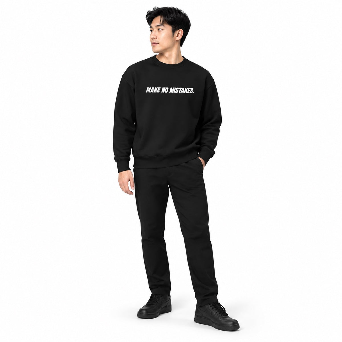 Full-body model wearing the black MAKE NO MISTAKES. sweatshirt with white chest print, black trousers and trainers, looking to the side on a white background.