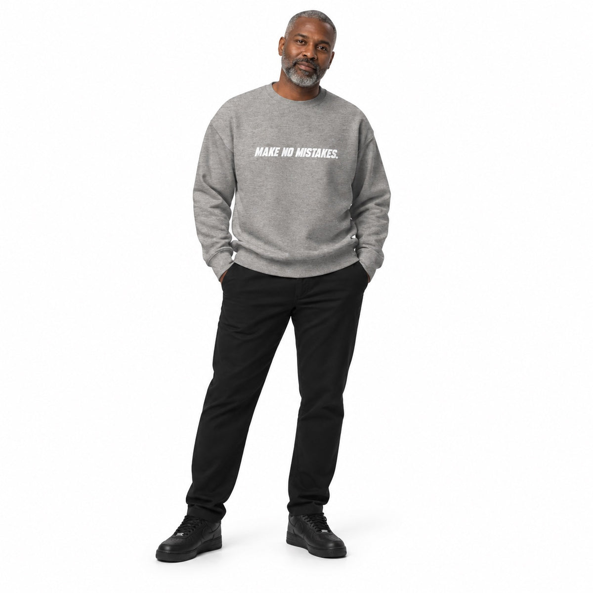 Full-body man wearing the carbon grey MAKE NO MISTAKES. sweatshirt with white chest print and black trainers, hands in trouser pockets on a white background.