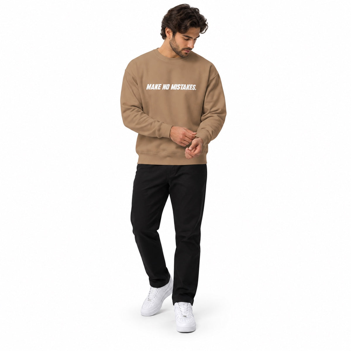 Full-body man wearing the latte MAKE NO MISTAKES. sweatshirt with white chest print, adjusting a sleeve cuff with white trainers on a white background.