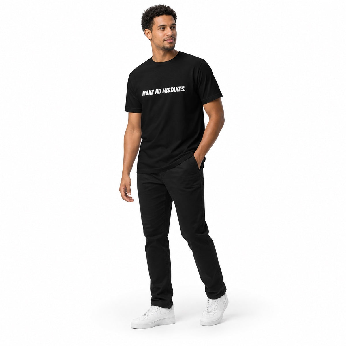 Full-body model wearing the black MAKE NO MISTAKES. T-shirt with white chest print, standing at an angle with one hand in a pocket and looking aside on a white background.