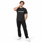 Full-body model wearing the dark grey MAKE NO MISTAKES. T-shirt with white chest print, smiling with one hand behind his neck on a white background.